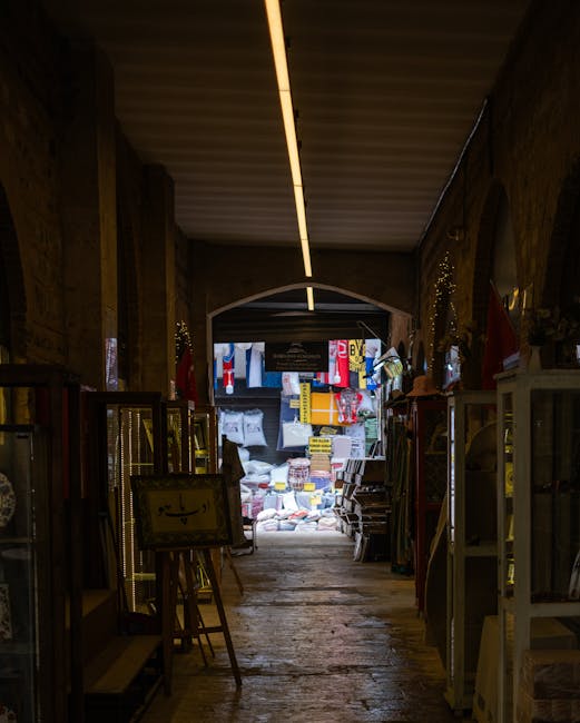 Inside a narrow retail space with wooden walls and ceiling, a long passageway leads to the exterior through an arched doorway. The interior is dimly lit, with a single strip light running along the ceiling. On each side of the corridor, there are glass display cabinets and shelving units filled with various small items, possibly ornaments or household goods, supported by metal frames. Near the entrance, stacks of cardboard boxes and wrapped packages are visible, indicating ongoing packing or relocation activities. Outside the doorway, bright light illuminates a street scene with colourful hanging banners, textiles, and miscellaneous goods, suggesting a busy market environment. The image captures the process of home relocation or moving logistics carried out by Lambeth Man and Van during a house removal project, highlighting the transition from interior packing to outdoor loading, with the focus on furniture transport and packing materials used in a typical narrow street move in Brixton.