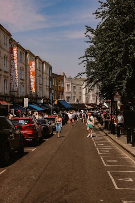 A busy street scene in Brixton Market with multiple parked cars along the left side, including a red vehicle and a black SUV, while pedestrians walk along the pavement and across the road. The street is lined with multi-storey terraced buildings featuring brick and painted facades, some with large banners or signage. On the right side, there is a row of metal bollards separating the pedestrian walkway, partially shaded by a large leafy tree. Visible are several cardboard boxes and wrapped furniture items indicating a home relocation process, with some being carried or prepared for loading. In the background, market stalls with striped awnings and more pedestrians browsing are visible. The scene is captured under a bright blue sky with a few clouds, conveying an active environment suitable for a furniture transport or moving service in the context of house removals in Lambeth, as supported by Lambeth Man and Van’s services.