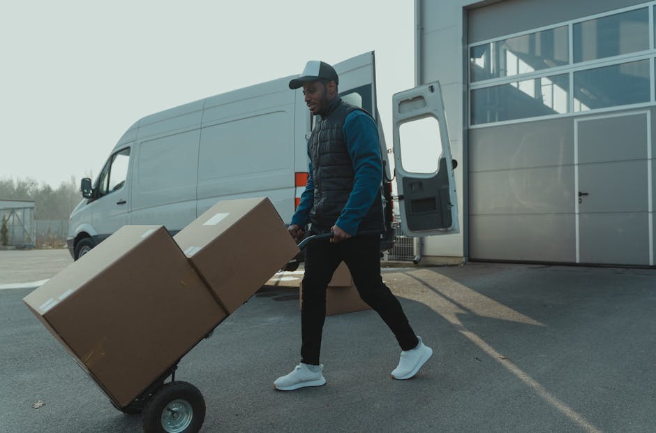 A man wearing a blue beanie, red and navy jacket, black trousers, and sneakers is loading cardboard boxes into the open rear cargo area of a silver van parked on a paved street. Several cardboard boxe