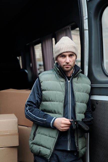 A young man with a beard and serious expression, wearing a black cap and white polo shirt, stands inside the open rear doors of a loaded moving van. The van's interior is filled with packing boxes mad